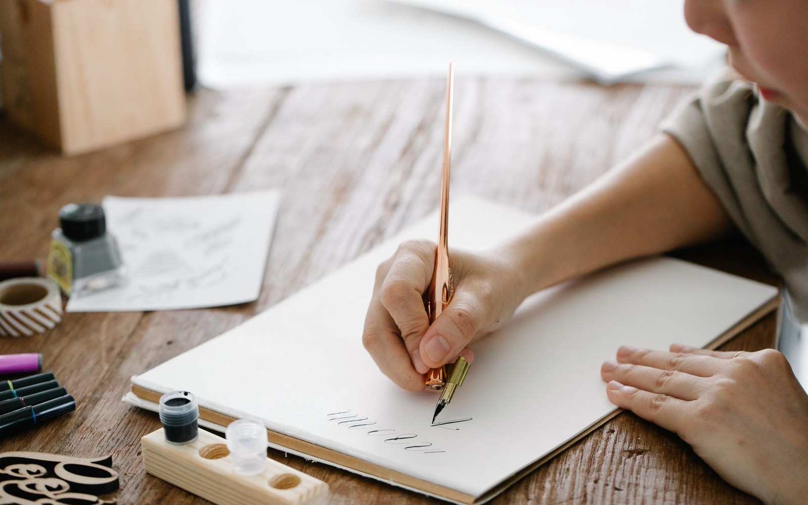 An adult woman's hands holding a nib over an ink drawing on a drawing board, a partially completed card face visible, photographed from above on a cloth-covered work surface